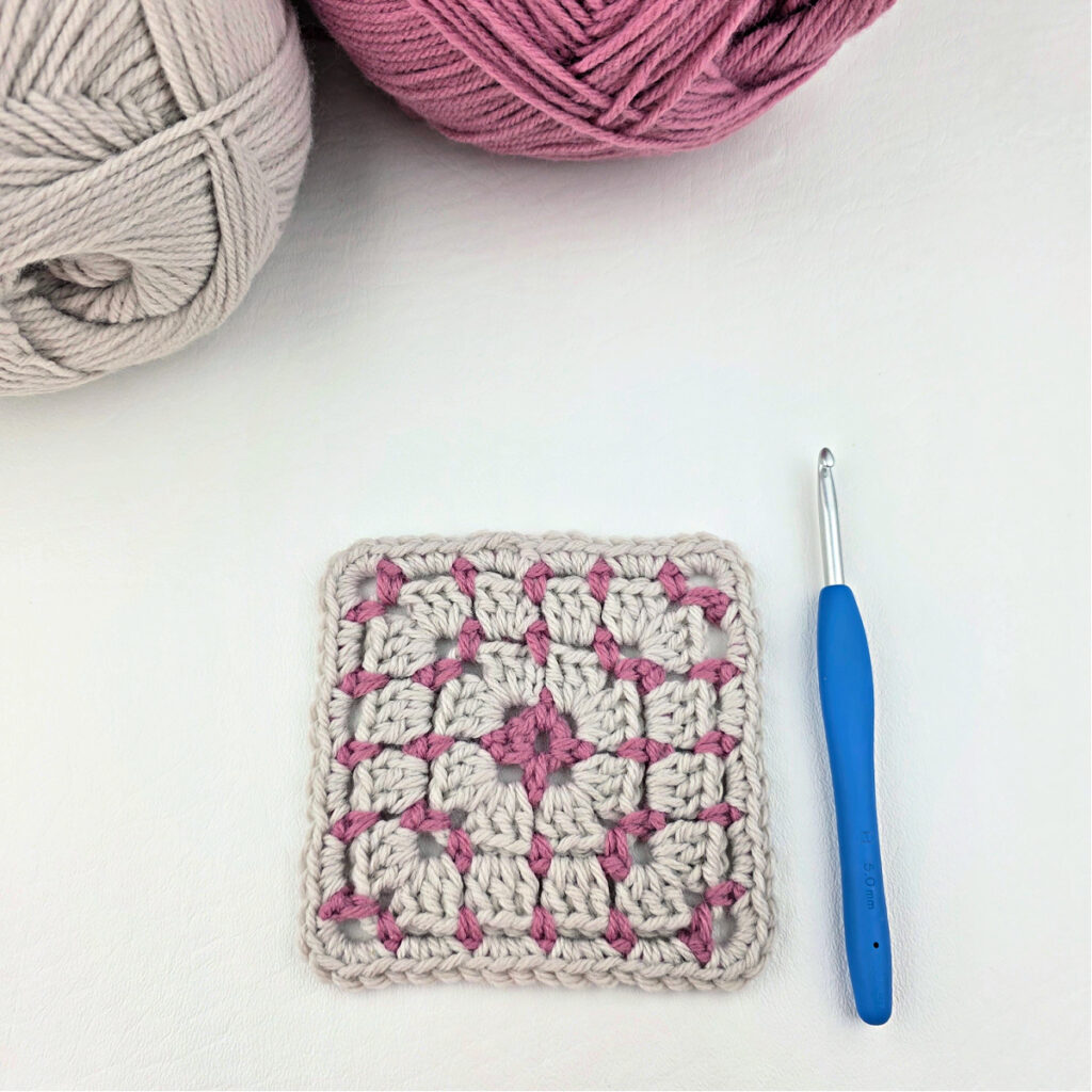 A white and pink block stitch granny square with two balls of yarn in the corner and a blue crochet hook.