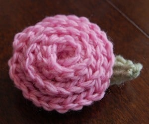 Pink crochet rose with small green leaf sitting on a wood table.