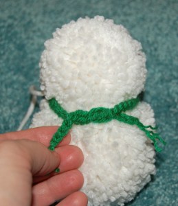 Small yarn pom snowman with green scarf being tied around its neck.