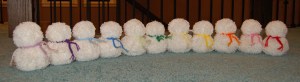 Four small yarn pom snowmen with scarves sitting on carpeted floor.