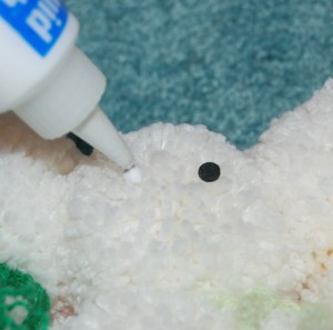 Black eyes being glued onto white yarn pom to make a snowman.