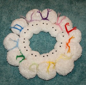 Snowman yarn pom wreath laying on floor with scarves attached.