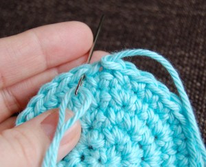 Hand holding small crochet circle and yarn needle to work an invisible join stitch.