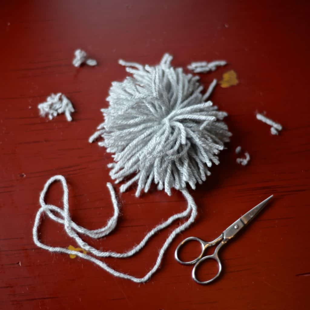 Grey yarn pom laying on red table with scissors.