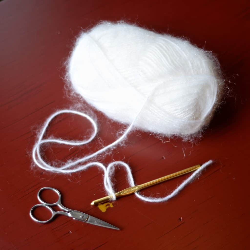 White yarn with halo laying with crochet hook and scissors on red table.