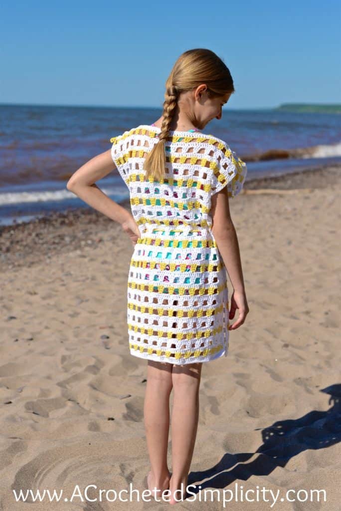 Girl modeling a white and yellow crochet beach cover up while standing on the sand at a beach.