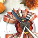 Crochet turkey napkin ring and flatware holder sitting on white dinner plate with fall flowers