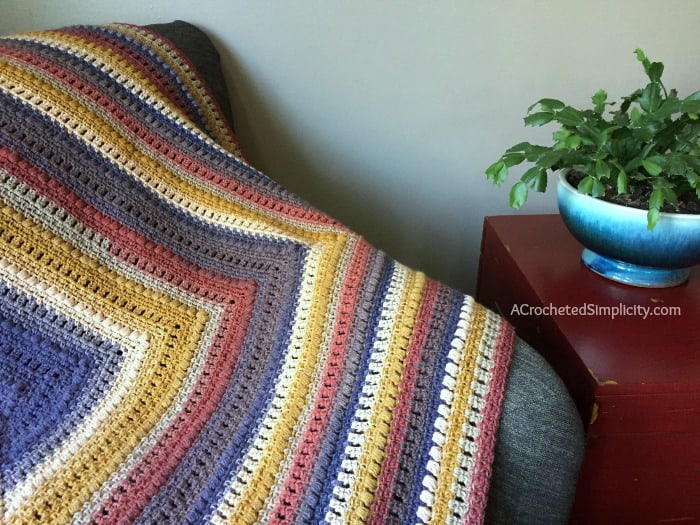 A crochet mandala blanket in warm tones spread out on a sofa next to a plant in a blue pot.