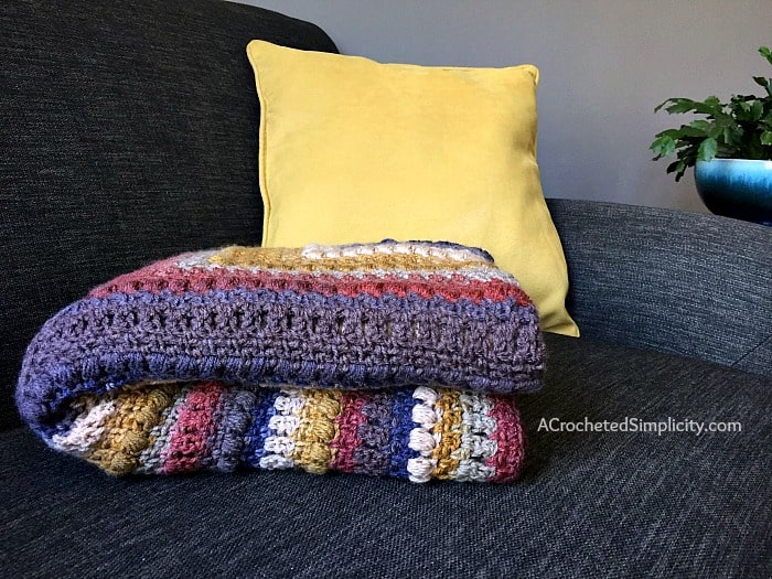 A crochet mandala blanket in warm tones folded on a sofa with a plant and yellow cushion next to it.