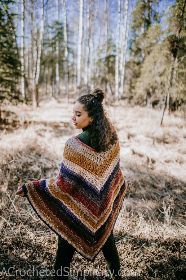 Girl standing with her back to the camera in a forest wearing a colorful crochet poncho.