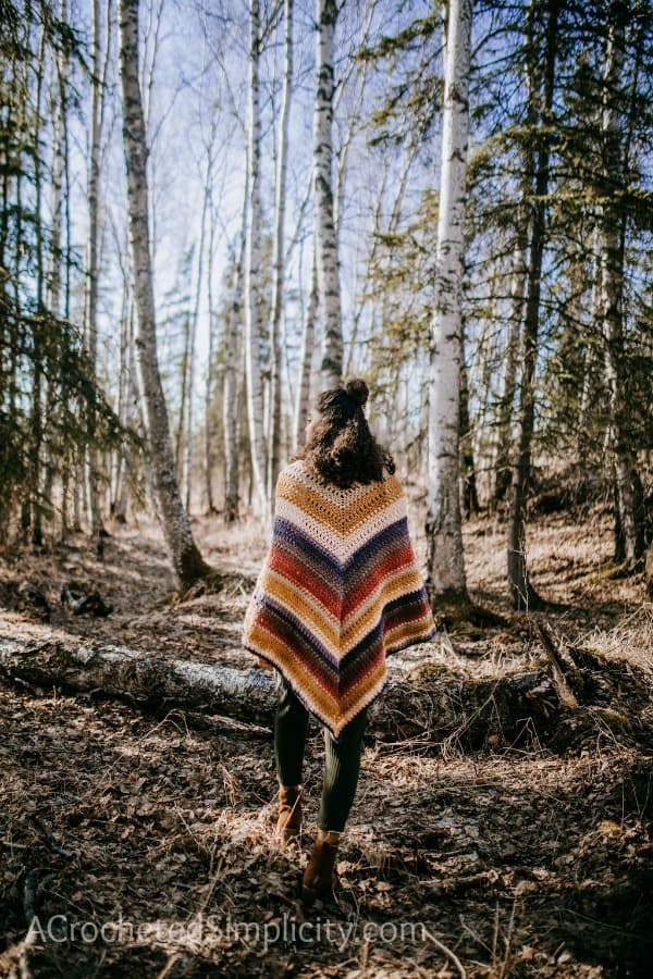 Girl standing with her back to the camera in a forest wearing a colorful crochet poncho.