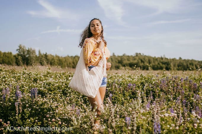 Young woman holding an ecru crochet tote bag on her shoulder standing in a field of lavender.