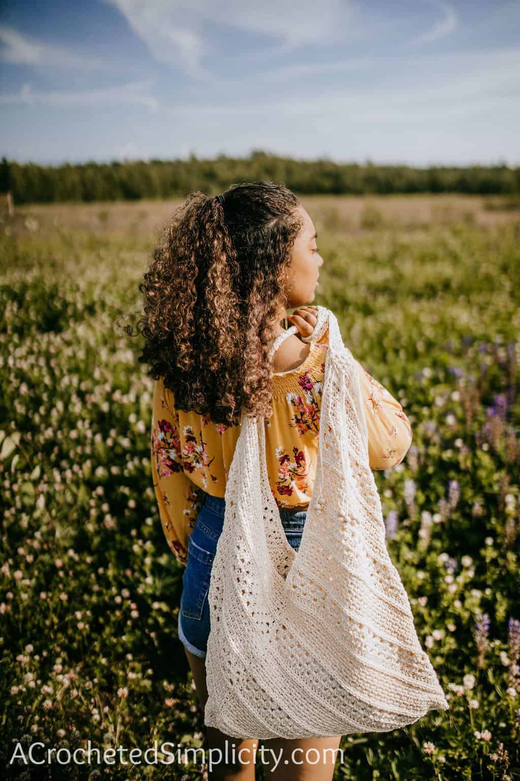 Young girl with dark curly hair holding a crochet tot bag over her shoulder while standing in a field of lavender.
