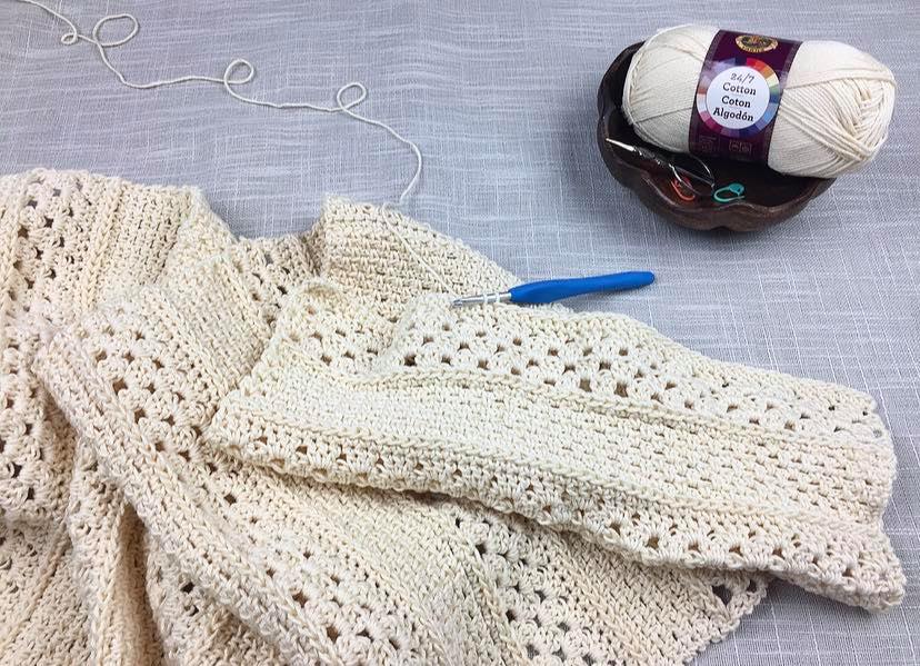 Ecru colored crochet rectangle with a blue crochet hook and small wooden yarn bowl.