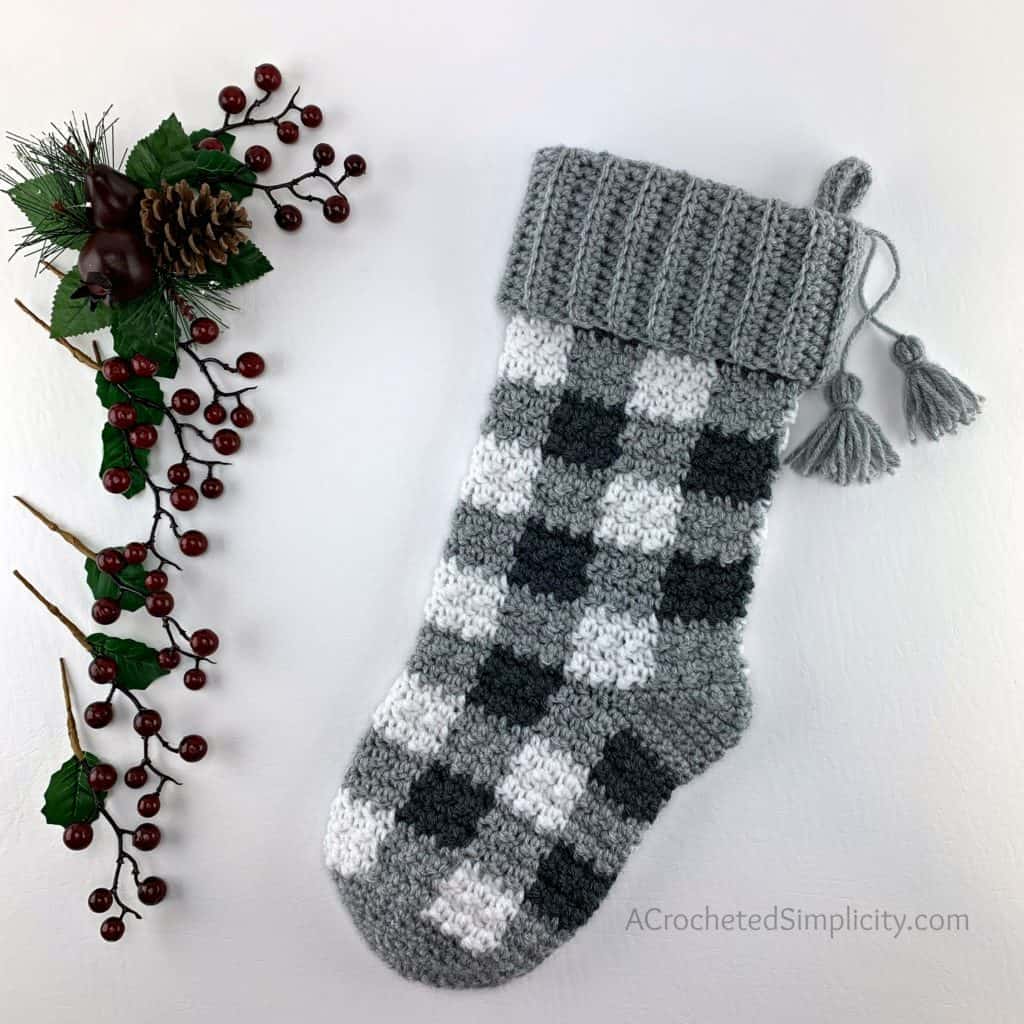 Buffalo Plaid Christmas Stocking in greys laying on white background with holly berries and pine cones