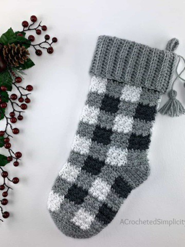 Buffalo Plaid Christmas Stocking in greys laying on white background with holly berries and pine cones