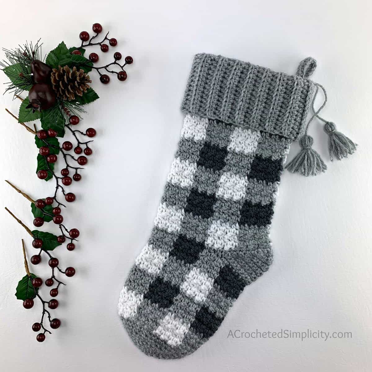 A black, medium grey, and white buffalo plaid christmas stocking laying next to holly berries.