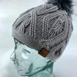 Grey crochet cabled beanie on glass mannequin head with dark grey faux fur pom.