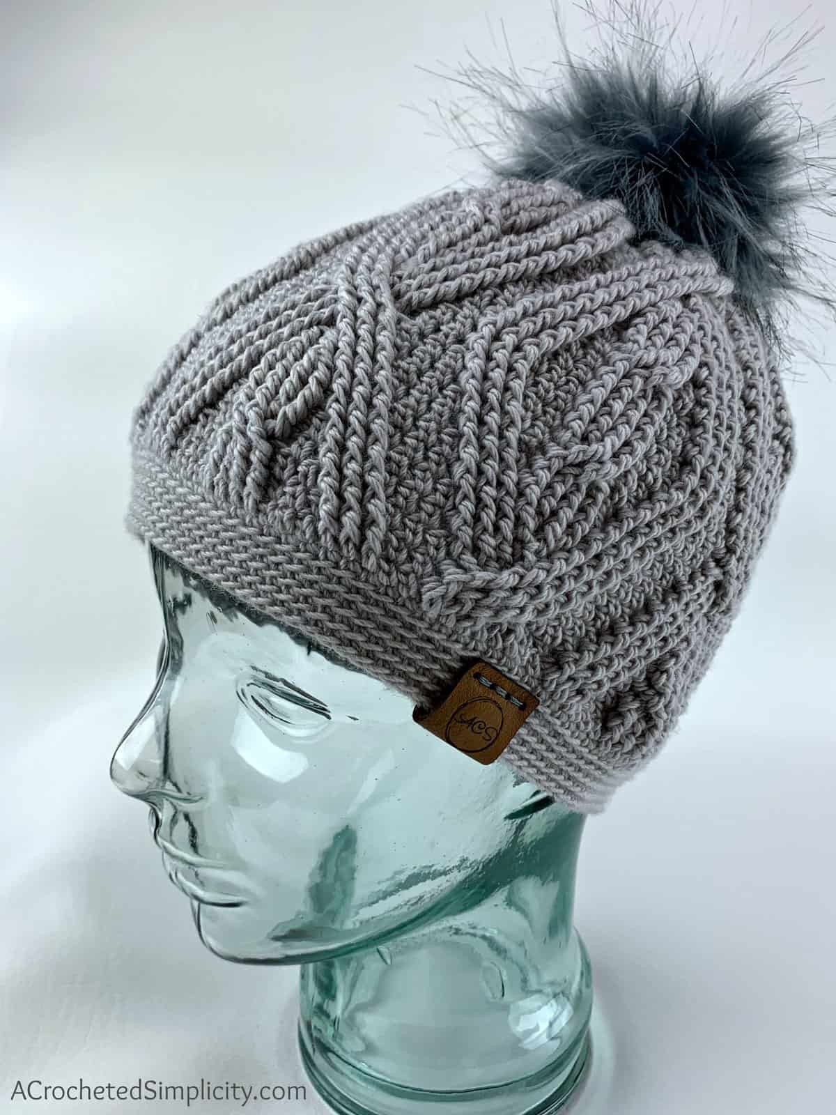 Grey crochet cabled beanie on glass mannequin head with dark grey faux fur pom.