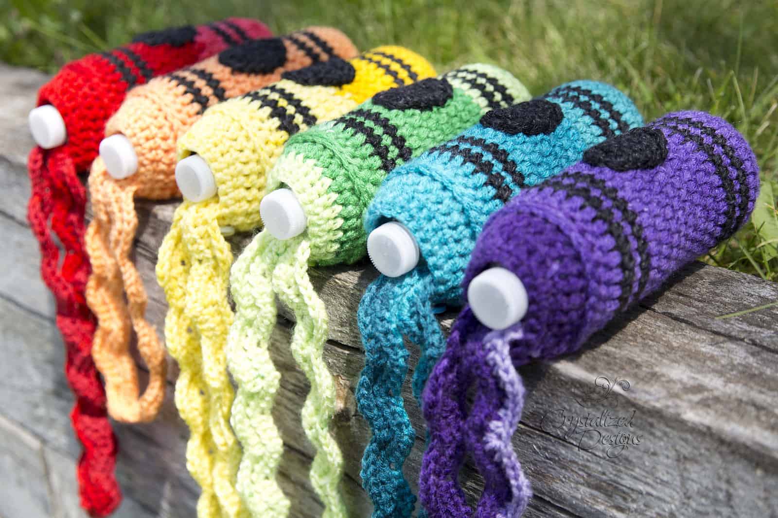 Rainbow colored crayon crochet water bottle holders laying on wooden fence.