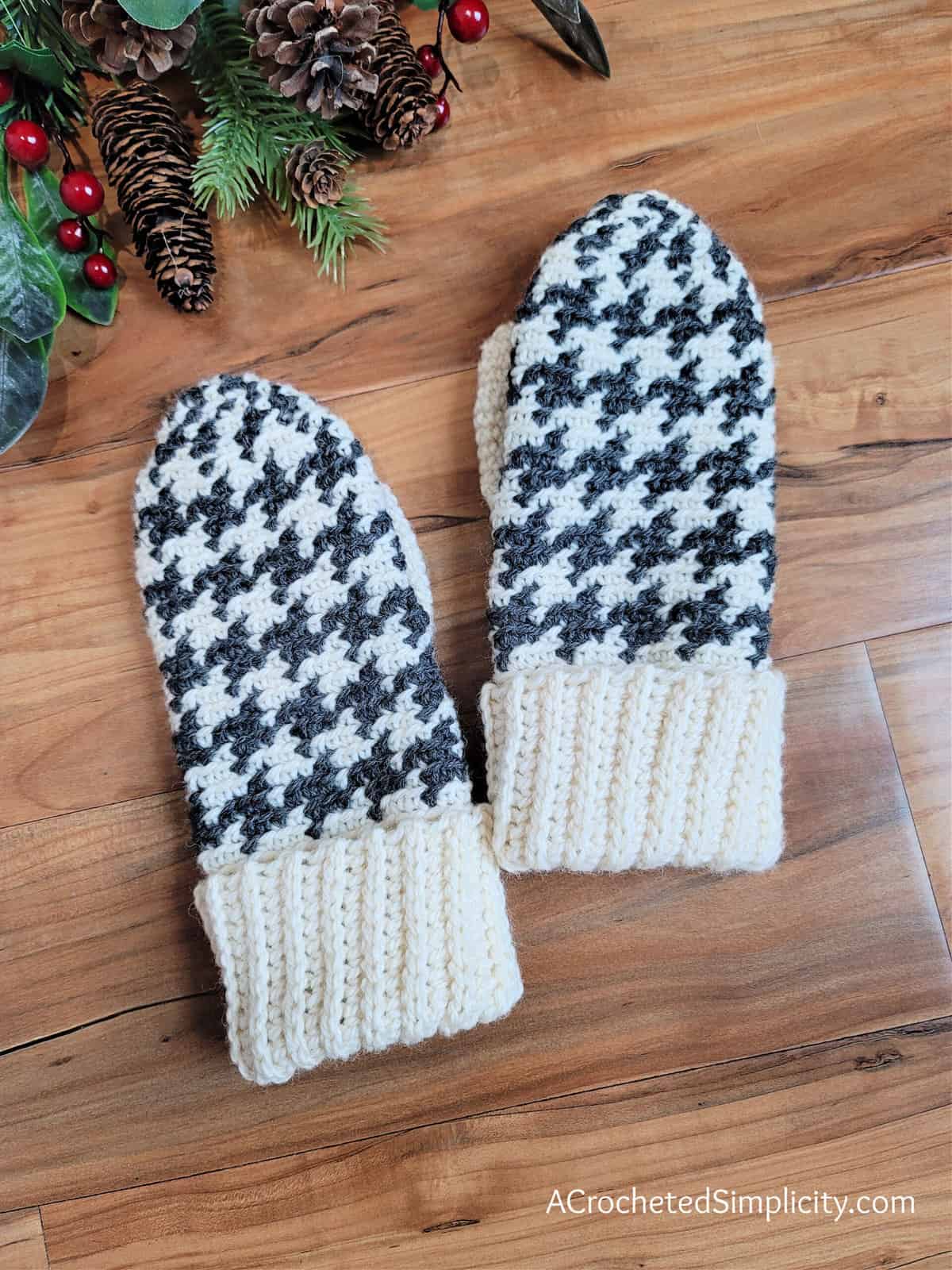 Two houndstooth crochet mittens laying on a wooden floor.