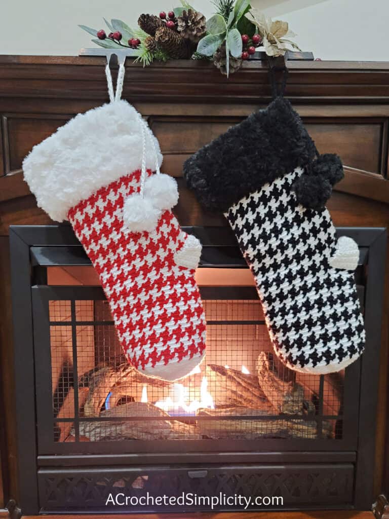 Two crochet houndstooth Christmas stockings with faux fur cuffs hanging on the front of a fireplace.