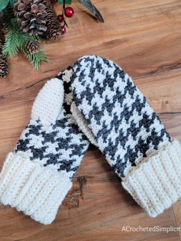 A pair of houndstooth crochet mittens laying on a wood floor with a holiday sprig of pine cones and berries.