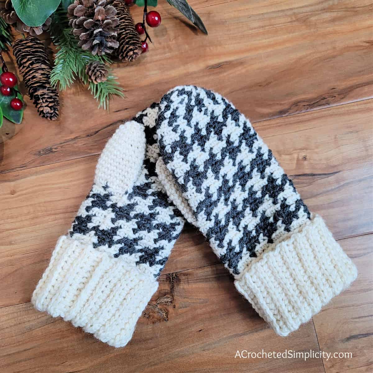 A pair of houndstooth crochet mittens laying on a wood floor with a holiday sprig of pine cones and berries.