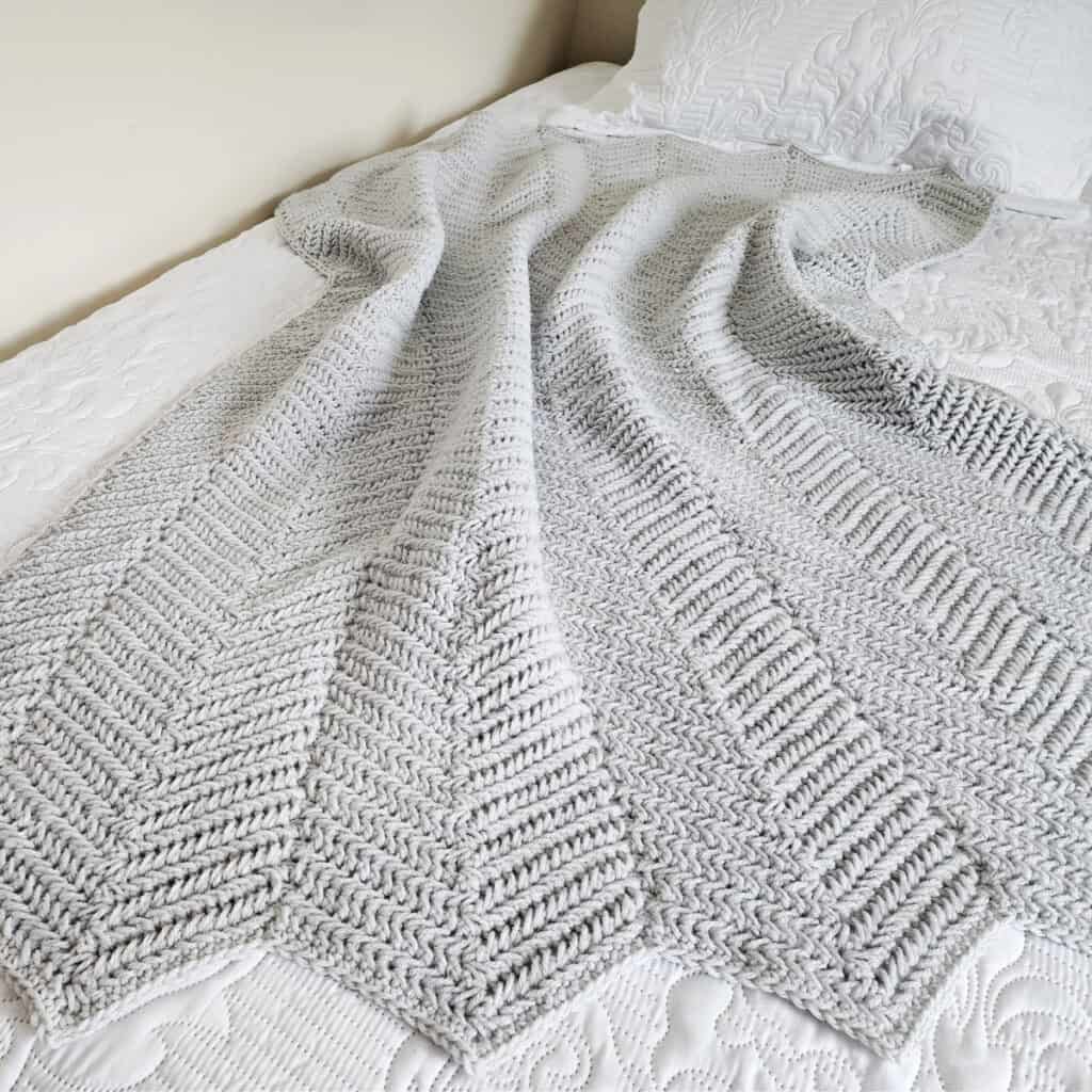Light grey crochet ripple blanket laying on bed.