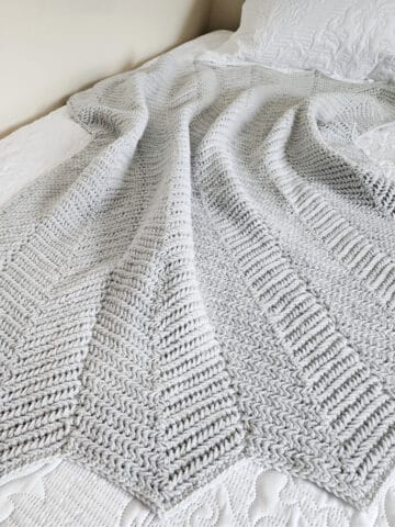 Light grey crochet ripple blanket laying on bed.