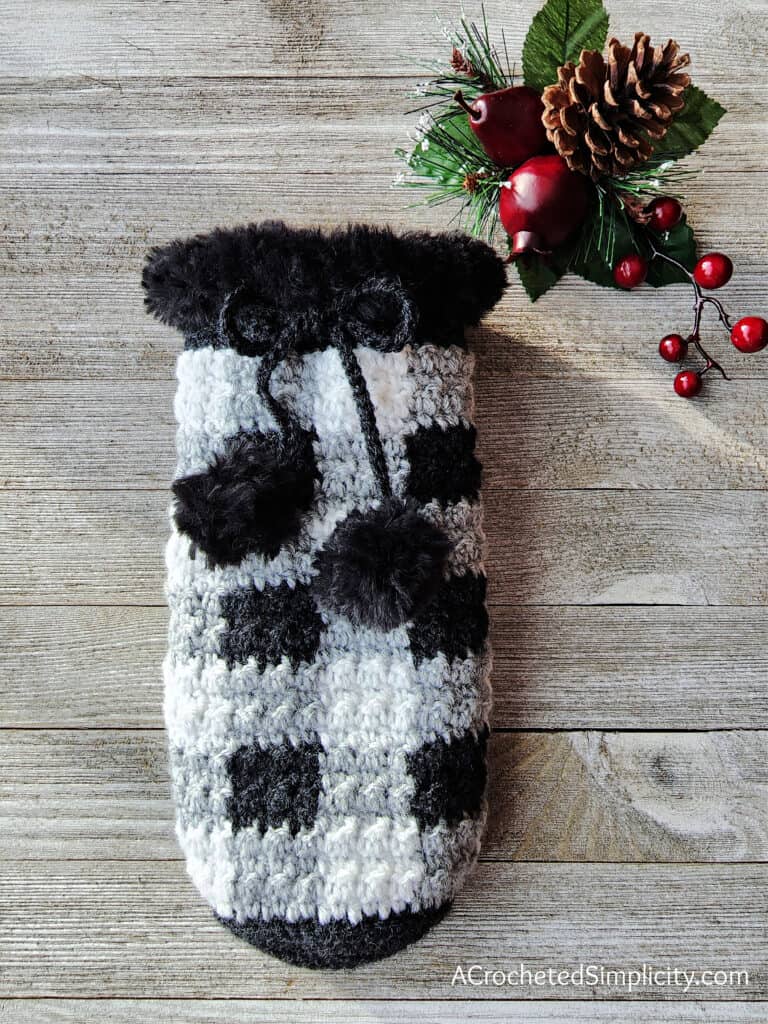 A white, black, and gray buffalo plaid crochet wine cozy laying flat on a wooden board.