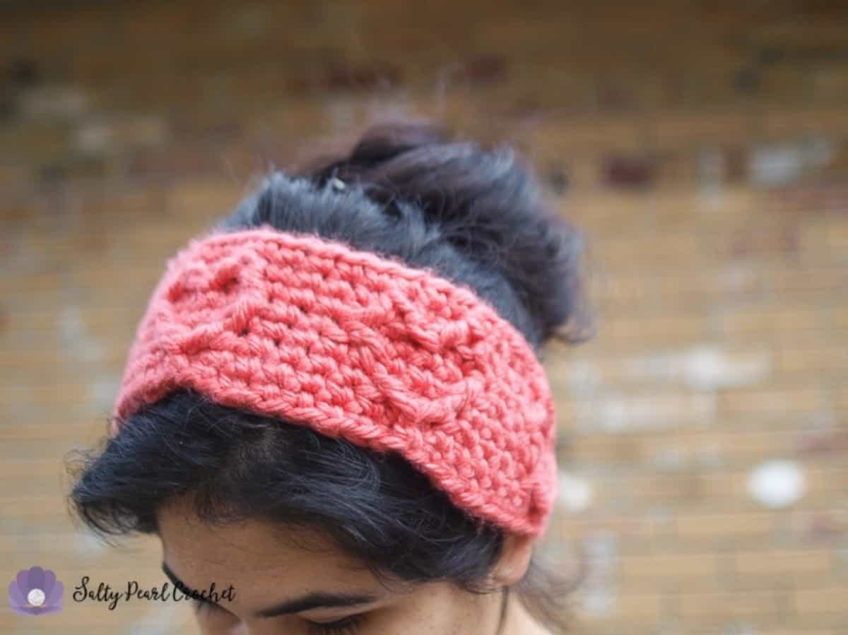 Woman modeling a medium pink, chunky yarn, crochet cabled heart ear warmer.