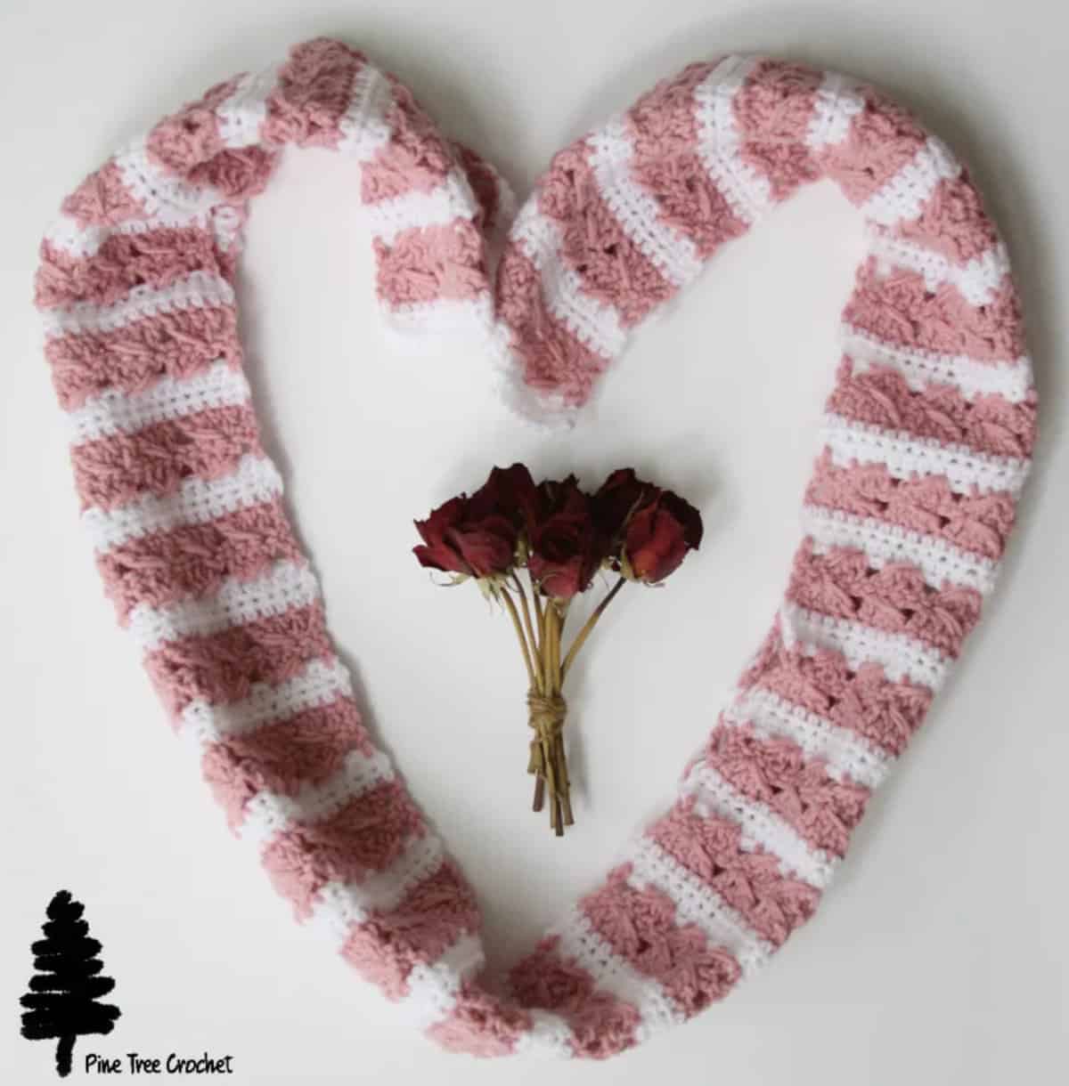 Pink and white striped crochet scarf posed in the shape of a heart with dried roses in the center.