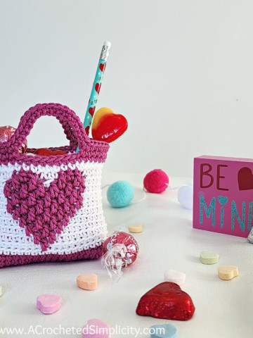 Mini crochet Valentine treat bag in white with a medium pink heart. Small candies scattered on the table.