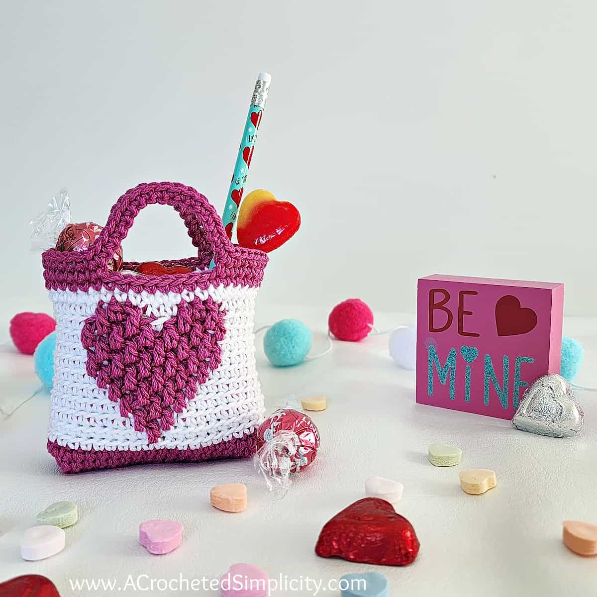 Mini crochet Valentine treat bag in white with a medium pink heart. Small candies scattered on the table.
