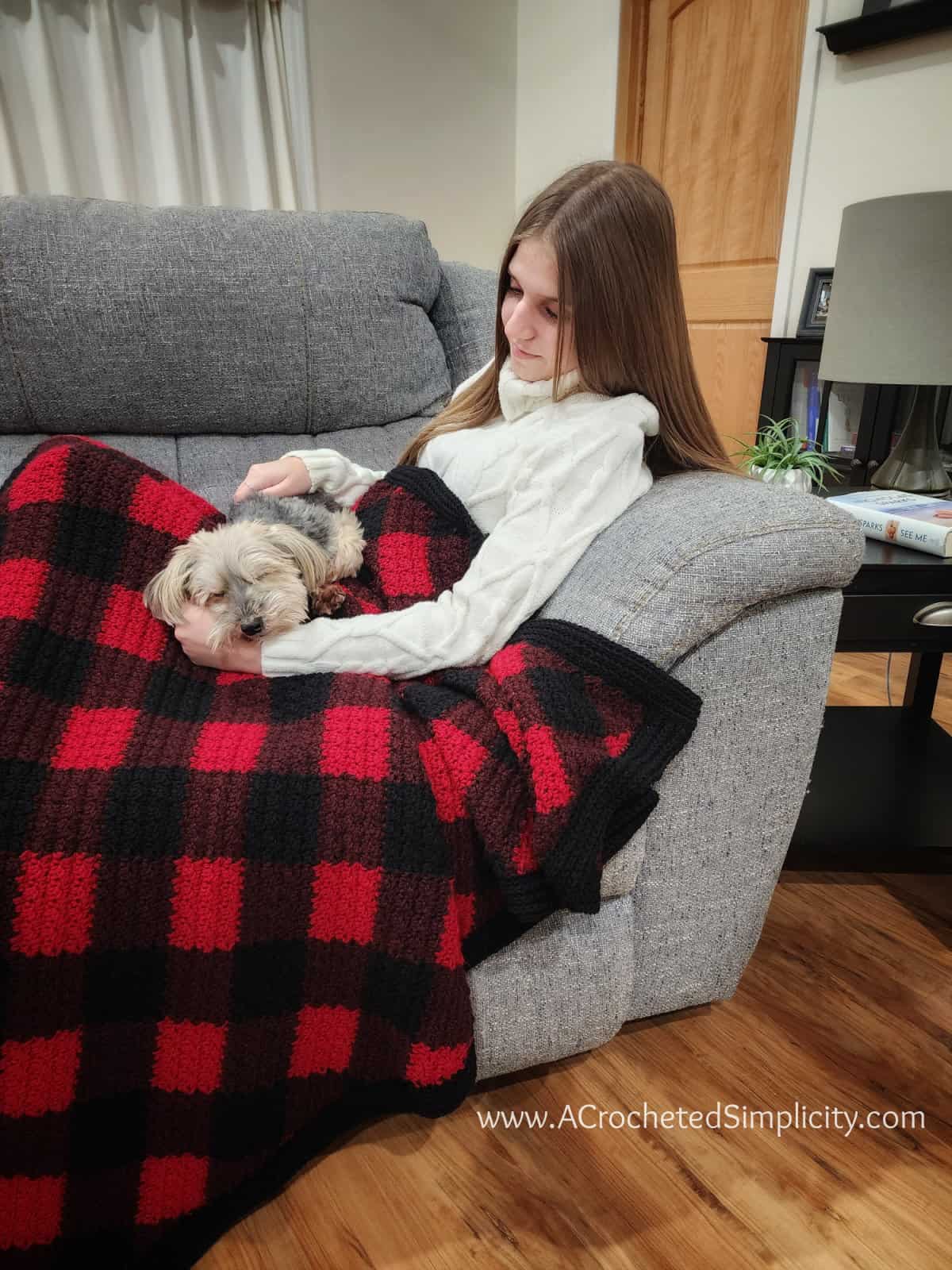 Girl sitting on a couch with a yorkie on her lap and a crochet buffalo plaid blanket.
