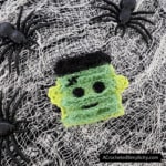 Crochet Frankenstein's monster made using cotton and scrubby yarns. Laying on netting with black plastic spiders.