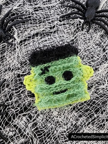 Crochet Frankenstein's monster made using cotton and scrubby yarns. Laying on netting with black plastic spiders.