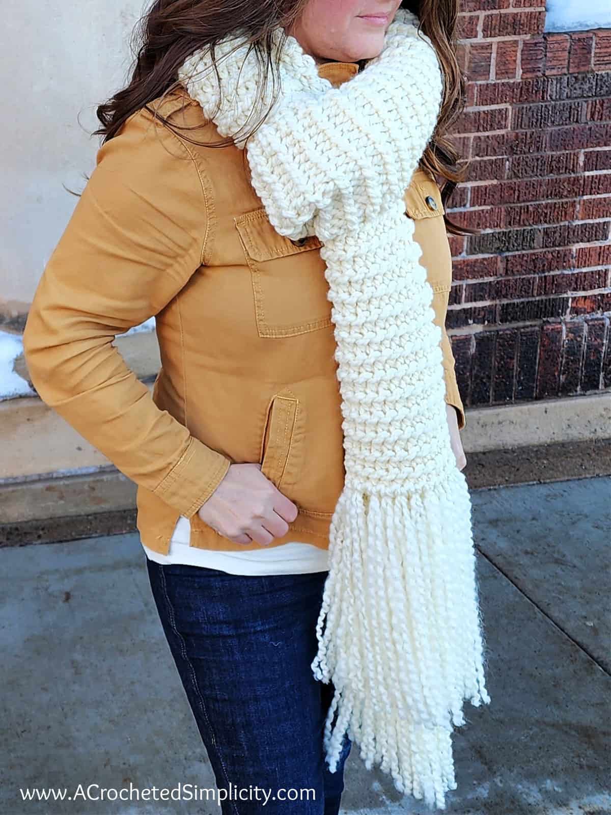 Woman wearing a cream colored knit look crochet scarf in front of brick building.