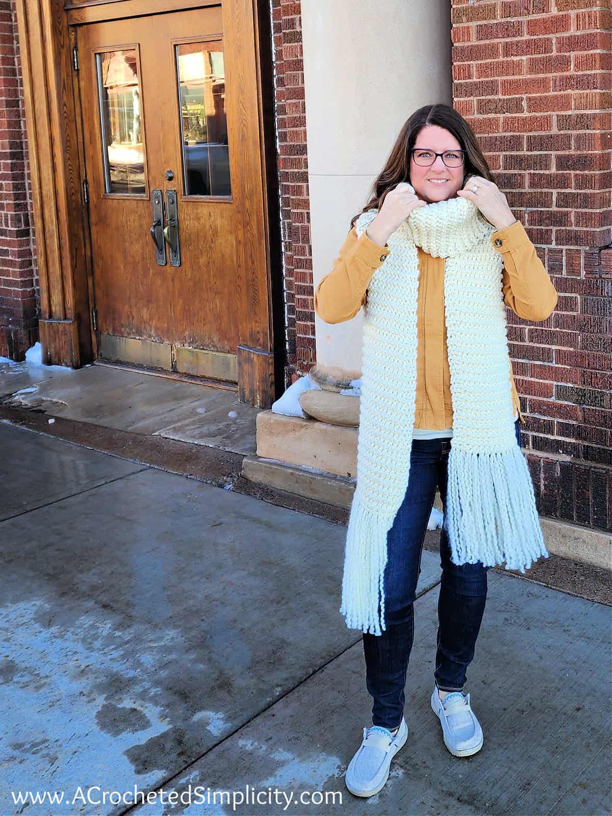 Woman modeling cream color knit look crochet scarf in front of a brick building.