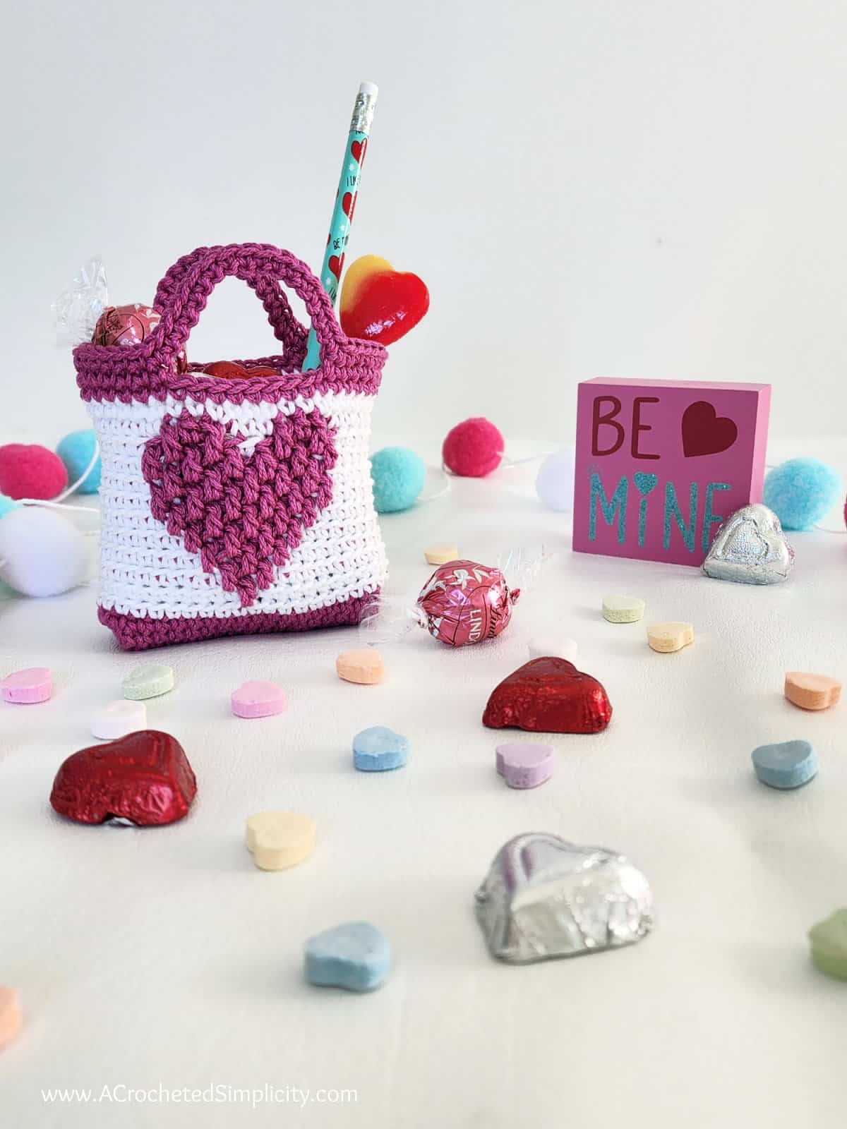 A small valentine treat bag in white and medium pink with candies scattered around it.
