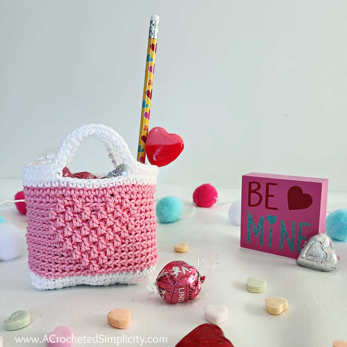 White and light pink crochet Valentine treat bag with a cute crochet heart. Truffles and chocolates are scattered around it.