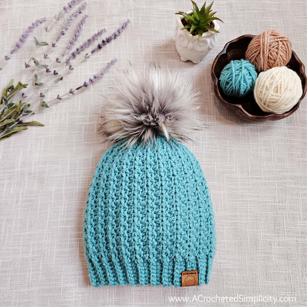 Aqua short row crochet hat with big fluffy faux fur pom, lavender and wooden yarn bowl.