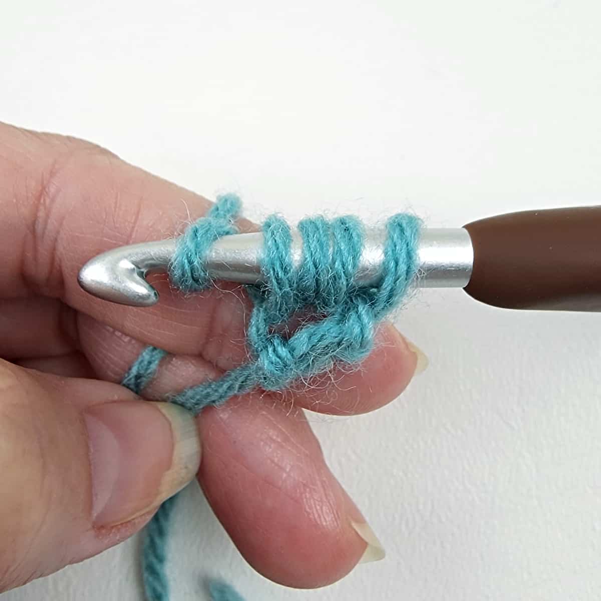 Hand holding blue green yarn and brown crochet hook.