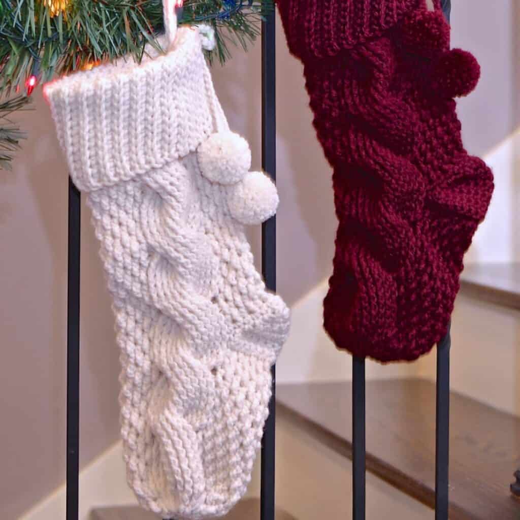Two crochet cabled Christmas stockings hanging on a staircase railing with garland.