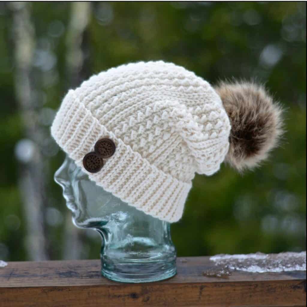 Fisherman color textured crochet beanie with a brown faux fur pom on a glass mannequin head.