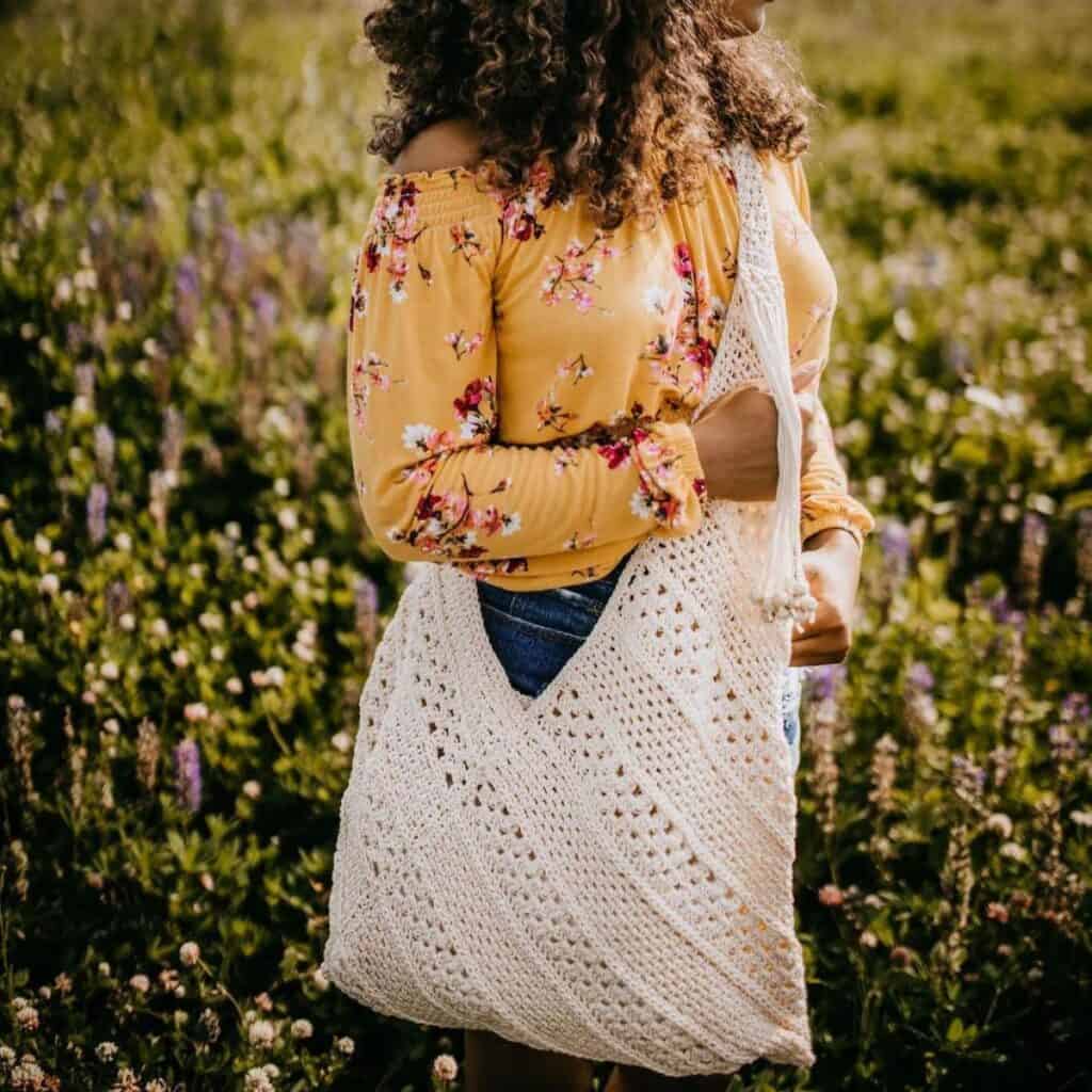 Young woman wearing a yellow floral blouse and cream color crochet shoulder bag in field of flowers.