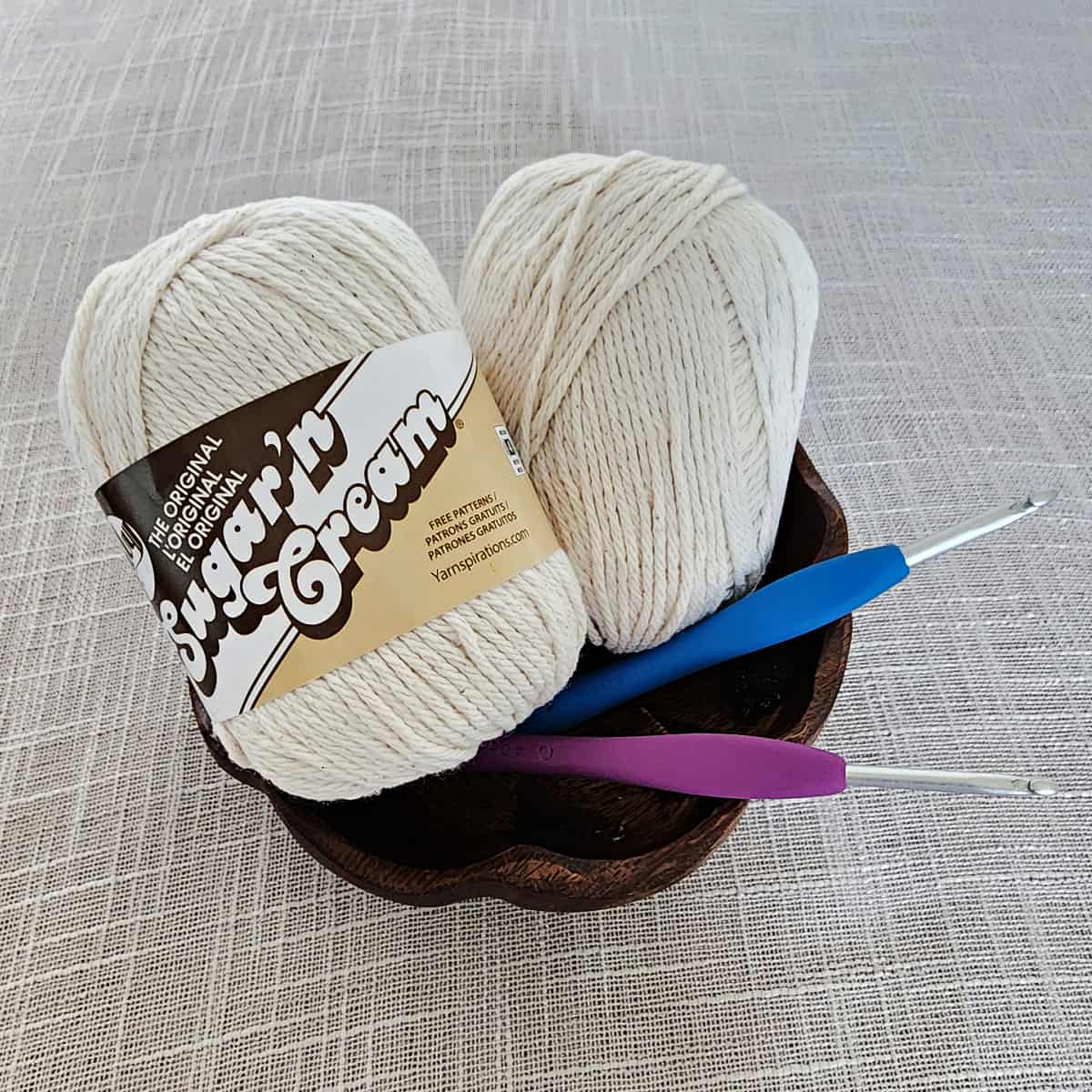 Two balls of ecru cotton yarn in wooden yarn bowl bolding crochet hooks.