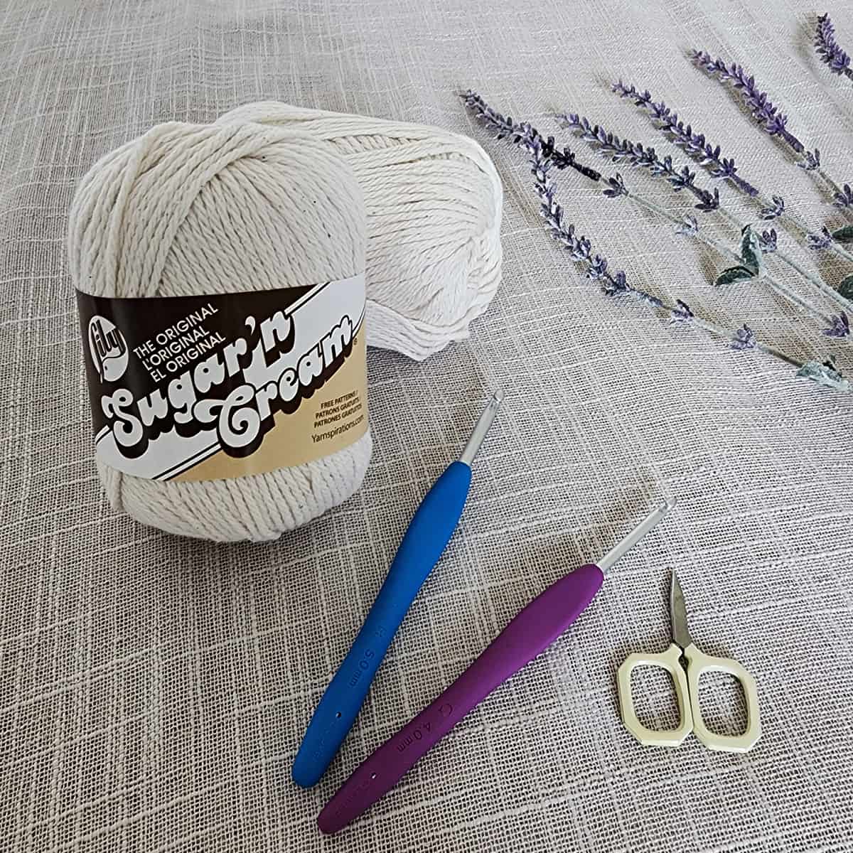 Two balls of ecru yarn laying on a cloth surface with crochet hooks, scissors, and a floral sprig.