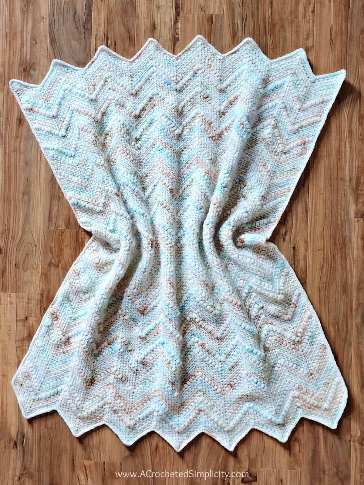 Crochet ripple beach blanket in light beachy colors laying on wood floor.